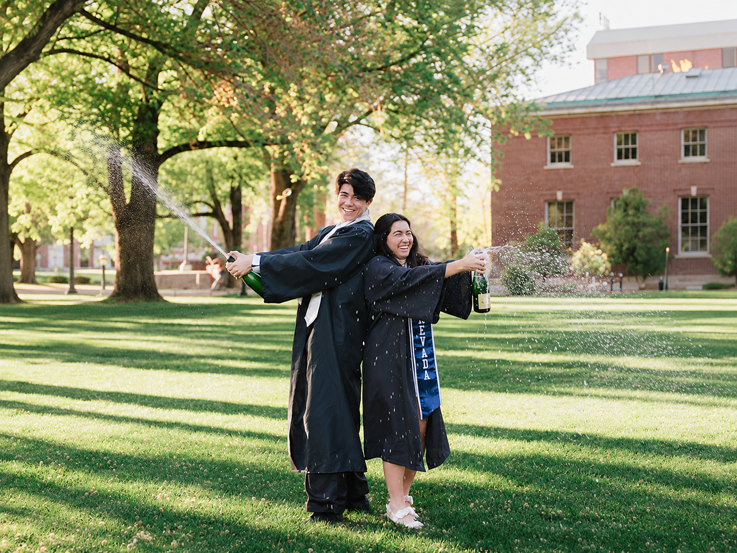 Two graduates celebrating with champagne on a campus lawn